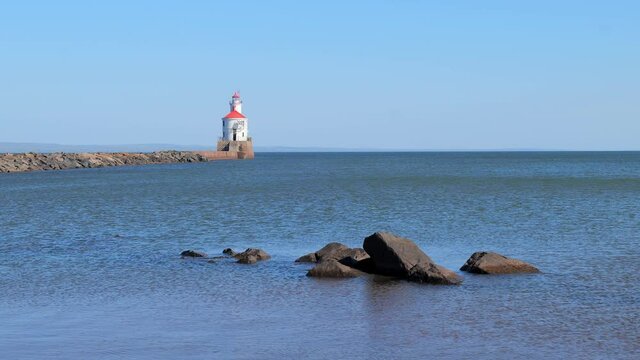 Lighthouse with red roof at the end of a pier on Lake Superior, and large boulders surrounded by ripples of blue water being blown away from the camera by the wind.