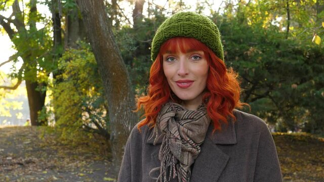 portrait of a bright girl with red hair and a green hat. looking at the camera and smiling against the backdrop of greenery in an autumn park