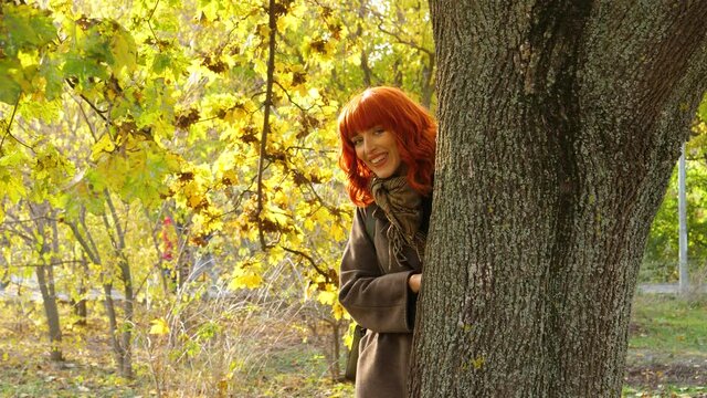 a girl with red hair in a coat, in an autumn park against a background of bright yellow foliage, looks out from behind a tree, smiles and then hides back
