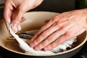 Closeup of Man hands put the fresh fish smelt in flour on frying pan. Unhealthy food on oil 