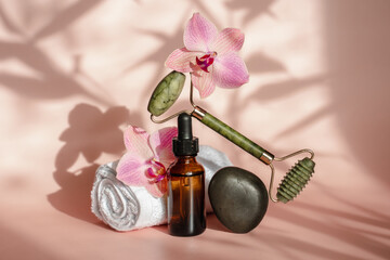 Photo for the spa salon. Roller and Guasha scraper for facial massage, massage oil, black hot spa stones, pink orchids, white towel on a pink background. 
Balance and levitation trending style