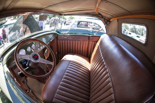 Interior View of Restored Vintage Automobile Dashboard Steering Wheel and Bench Style Seat, Out of Focus People and Autos in Background