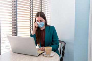 Young woman in suite with protective mask is working on computer and talking on mobile phone.