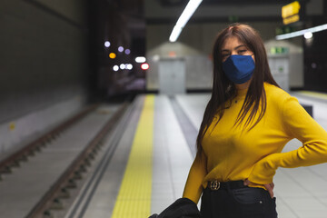 joven y atractiva muchacha con camisa amarilla y mascarilla azul para el coronavirus espera el metro en la parada con paciencia. - obrazy, fototapety, plakaty