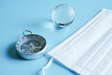 青い紙の上に置いた不織布マスクと方位磁石とガラスの地球。コロナ禍の世界はどの方向に進むのか。
