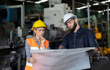 Male and female mechanical engineers or factory workers with safety gears and hard hats discuss about production project using blueprint plan in a warehouse