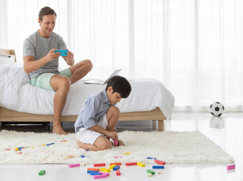 Father using smartphone to take photos of his mixed-race son who playing with a colorful wooden block in home bedroom. Idea for lifestyle of working from home