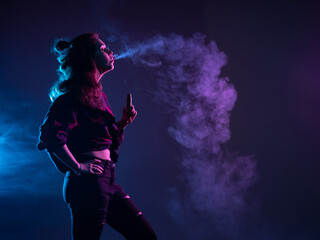 A girl with a VAPE stands in profile. A woman smokes a VAPE and lets out smoke. Girl vaper on a dark background. A girl with an e-cigarette looks at the smoke. Vaping concept with place for text.
