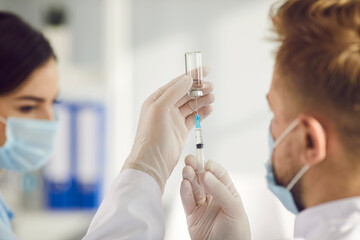 Close-up of filling syringe with vaccine from ampule in man doctors hands before vaccinating young woman against 19-ncov infection during pandemic times in medical clinic office.