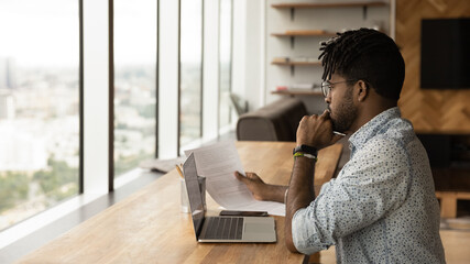 Concentrated young african man read printed document before laptop screen compare data think on...