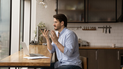 Inspired male remote worker sit at home office in kitchen before laptop pc dictate audio message via voice app on cell. Young man freelancer ask mobile phone ai assistant to search information online