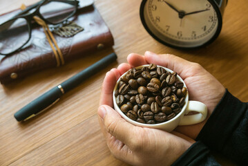 Selective focus of a hand holding a cup full with coffee beans ,pen, notebook, glasses and alarm...