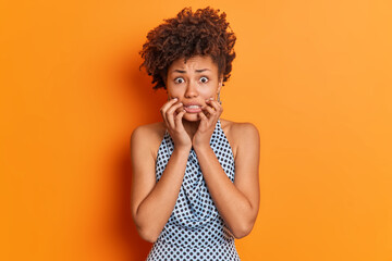 Nervous worried Afro American woman clenches teeth looks embarrassed at camera wears fashiobnable outfit afraids of bad consquences poses in studio against orange background. Scared female model - obrazy, fototapety, plakaty