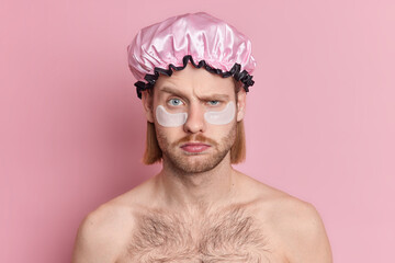 Portrait of displeased European man raises eyebrows looks with grumpy expression applies collagen patches under eyes to reduce wrinkles and fine lines stands shirtless wears bath hat isolated on pink