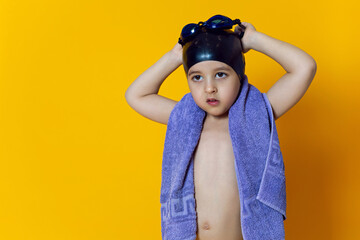 child athlete wearing blue swimming goggles and a black rubber cap stands in the studio on a yellow background