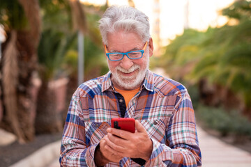 Smiling senior man using technology with smartphone for message. Standing outdoor in a public park at sunset. A white-haired people with beard and glasses