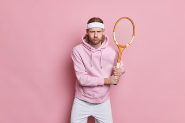 Serious active sportsman stands with tennis racket plays favorite game goes in for sport for health dressed in active wear enjoys active leisure isolated over pink background challenging himeself