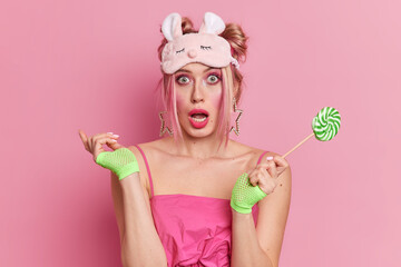 Obraz na płótnie Canvas Studio shot of surprised blonde young woman with vivid makeup keeps mouth opened wears sleepmask and dress holds lollipop isolated over pink background. Shocked beautiful female with sweet candy
