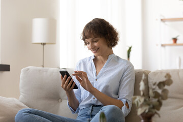 Smiling young woman rest on sofa at home look at smartphone screen shopping on internet on gadget. Happy Caucasian female renter relax on cozy couch in living room use cellphone, browsing web.