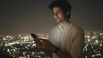 shot of a young handsome Indian male sitting on a terrace of building having a virtual conversation through text message using a mobile phone against the illuminated cityscape at night - Powered by Adobe