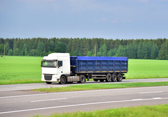 Truck with tipper semi trailer driving along highway on the green agriculture field background. Modern Dump Semi-Trailer Rear Tipper Truck Trailer. Goods Delivery and Services and Transport logistics