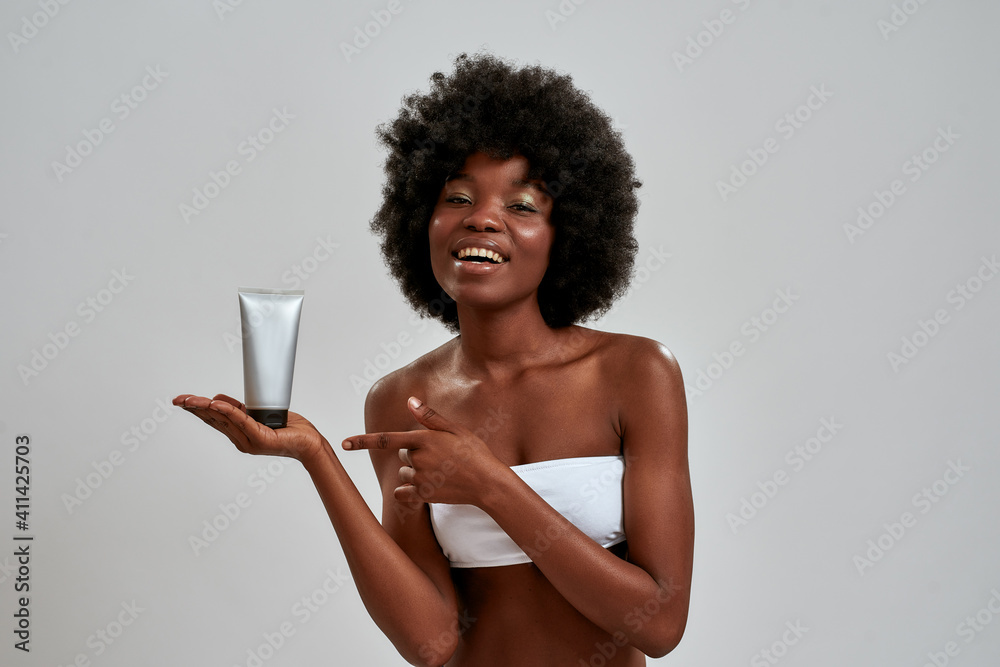 Wall mural Joyful young half naked african american woman holding beauty product bottle and pointing at it, smiling at camera isolated over gray background