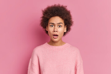 Photo of puzzled surprised African American woman stares at camera with great wonder and disbelief keeps mouth opened dressed in casual jumper isolated over pink background. Face expressions