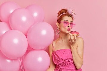 Attractive woman dressed in nineties style keeps lips folded holds appetizing doughut near mouth wears sunglasses and dress holds inflated balloons celebrates festive occasion isolated on pink wall