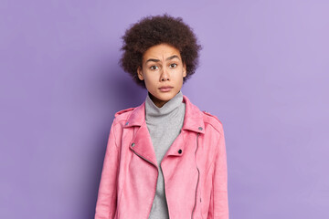 Fototapeta na wymiar Puzzled beautiful young African American woman in fashionable pink jacket reacts on something with dissatisfied expression has natural curly bushy hair isolated over vivid purple background.