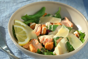 Caesar-style salad with roasted salmon pieces, croutons, green lettuce leaves, slices of lemon and Parmesan cheese, under sauce in a bowl. Top view