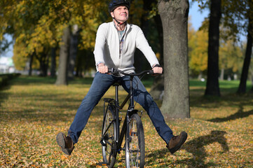 Happy mature Caucasian man in casual clothing and bike helmet with raised feet on his city bicycle looking upward in an autumn city park