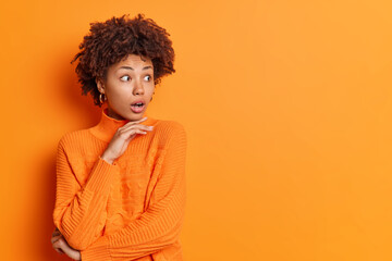 Studio shot of stupefied African American woman holds chin and stares aside dressed in casual orange jumper reacts on astonishing relevation poses indoor blank copy space for your advertisement