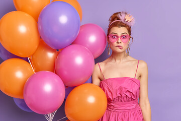 Obraz premium Puzzled woman with surprised face expression stares at camera wears crown pink heart shaped sunglasses and dress being on theme party holds inflated colorful balloons isolated over purple wall