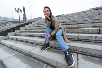 A young woman in a hooded beige coat and black sneakers sits on a grey marble staircase in clear weather. High quality photo