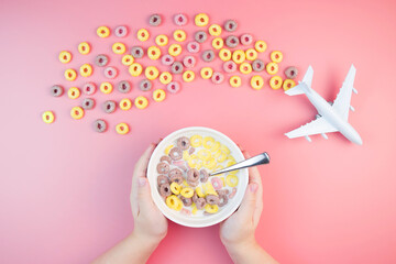 Colorful fruit corn rings with milk in the bowl, children's hands, airplane on a pink background. Children's breakfast option with cornflakes.