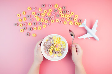 Colorful fruit corn rings with milk in the bowl, children's hands, airplane on a pink background. Children's breakfast option with cornflakes.