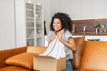 Overjoyed young African-American woman unpacking cardboard parcel box at home, cheerful girl hugs...