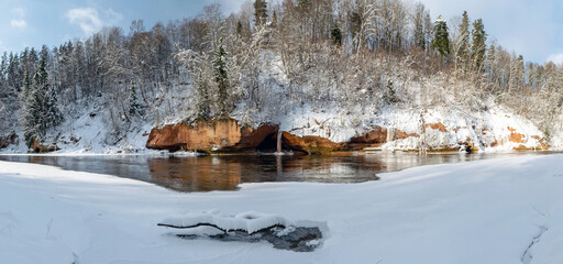 panoramic winter landscape with snowy sandstone cliff and frozen ice falls, slow river water, snowy trees on the river bank, Latvia