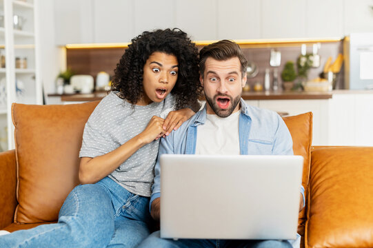 Shocked multiracial couple looks at the laptop screen with opened mouths, an African woman and caucasian guy surprised with unexpected new, watching a scary movie sitting on the couch at home