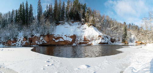 panoramic winter landscape with snowy sandstone cliff and frozen ice falls, slow river water, snowy trees on the river bank, Latvia