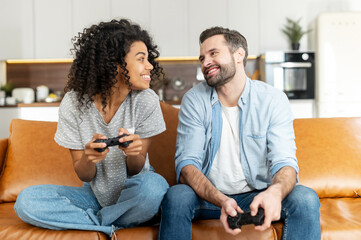 Excited multiracial couple enjoys playing video game, an african woman and a caucasian man hold wireless joysticks look at each other ready to fight. Interracial couple spends leisure time at home