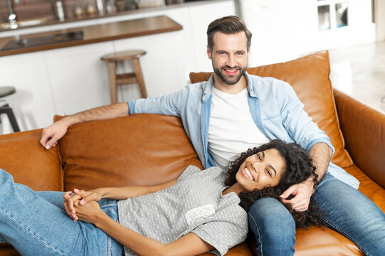 Smiling multi-ethnic couple in love rests together at home, spends a lazy weekend on the couch in cozy living room. African woman lies on the laps of her boyfriend in relaxed pose, looks at camera