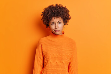 Fototapeta na wymiar Horizontal shot of displeased young African American woman frowns face looks angrily at camera dressed in casual jumper isolated over bright vivid orange background. Monochrome. Emotions concept