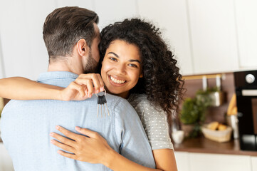 Charming and glad African-American woman holds keys in embraces of a guy. Happy interracial couple moved in new house, buying a real estate