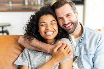 Lovely multiracial couple in embraces in cozy living room at home. A smiling caucasian guy is hugging charming african girl. Love and affection concept