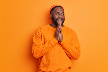 Positive dark skinned bearded man smiles happily keeps palms together has belief in better wears casual long sleeved jumper and hat poses against vivid orange background. Happy emotions concept