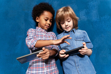Happy multiracial  children using applications on smartphones. Little european kid boy with african american foster brother playing online on phones together on blue studio background.