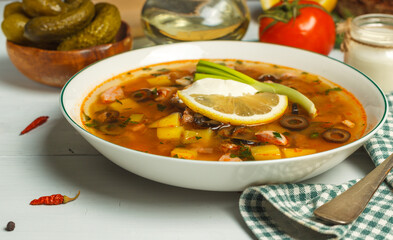 Solyanka, meat soup with pickles and olives, in a white plate on a light wooden table, close-up