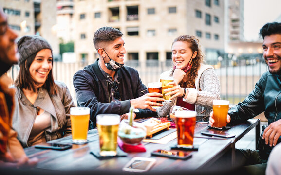 Young friends drinking beer with open face mask - New normal lifestyle concept with milenials having fun together talking at outside brewery bar - Warm filter with focus on left central guy