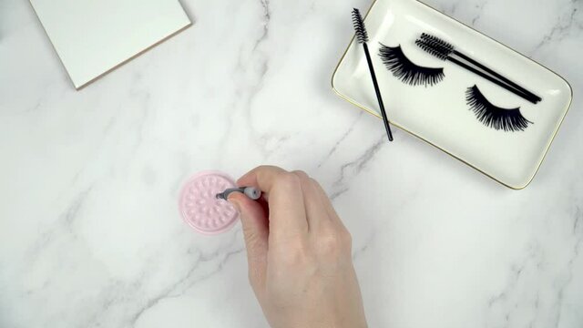 Eyelash extension glue being dispended into pink holder. Professional how to dispense glue for lash extensions beauty treatment set against a white marble background with text space